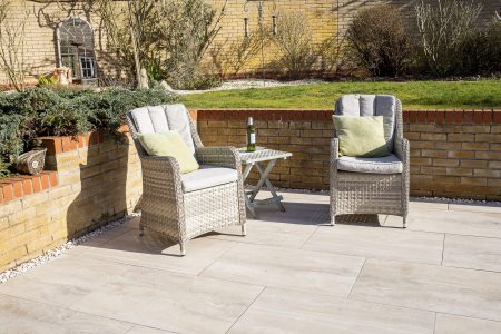 Paved garden patio with two wicker armchairs, cushions and side table on porcelain slabs, brick wall and planting behind.