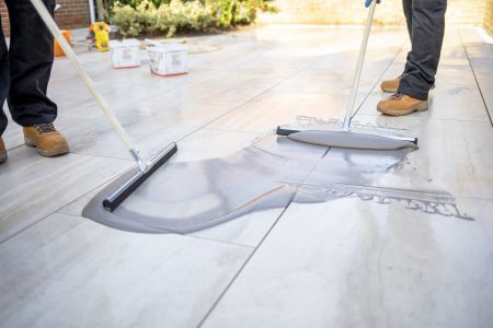 Porcelain paving installation with jointing tool and grout being applied between grey stone-effect tiles on a patio