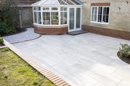 Light grey porcelain paved patio with brick edging adjoining brick house extension with conservatory.