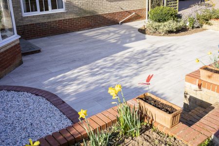 Porcelain paved patio with brick edging, adjacent pool with yellow flowers and ornamental grasses in borders.