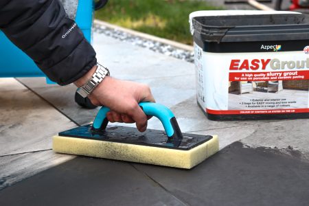Person applying grey grout to paving with blue grouting tool and yellow sponge, EASYGrout product visible.